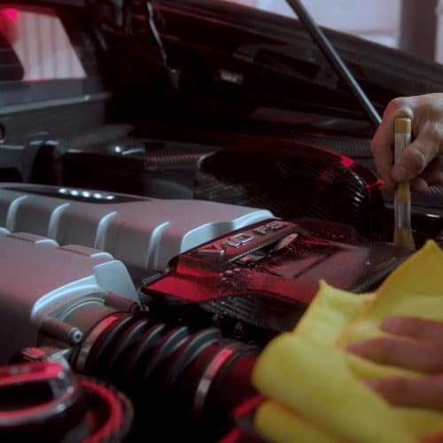 Detailing technician cleaning the engine bay of a luxury car with a brush